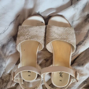Beige sandaler med spets och korksula från Zoey - Säljer ett par beige sandaler från Zoey med bred rem i spets och justerbart spänne runt vristen. Skorna har en bekväm plattformssula i kork och öppen tå. Perfekta för varma dagar och ger en somrig känsla till outfiten.