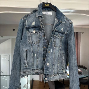 Blå jeansjacka från Z1975 Denim - Säljer en klassisk blå jeansjacka från Z1975 Denim med slitna detaljer och silverfärgade knappar. Jackan har två bröstfickor med lock och knapp samt två sidofickor. Perfekt för dig som gillar en avslappnad och trendig stil.