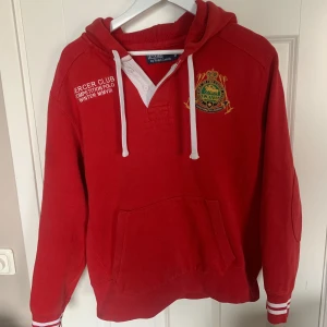Röd hoodie från Polo Ralph Lauren - Röd hoodie från Polo Ralph Lauren med broderad logga och text på bröstet. Tröjan har vit knappslå, huva med vita snören och ribbade muddar med vita detaljer vid ärmslut och nederkant. Perfekt för en sportig och avslappnad stil.