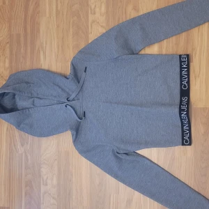 Grå hoodie från Calvin Klein Jeans - Säljer en stilren grå hoodie från Calvin Klein Jeans med svart resår och vit logga längst ner. Tröjan har huva med dragsko och långa ärmar. Perfekt för en avslappnad och trendig look.