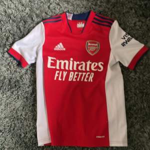 Säljer en Arsenal hemmatröja från säsongen 2021/22. Tröjan är röd med vita ärmar och blå detaljer, har klubbmärke och Adidas-logga på bröstet samt sponsortryck 'Emirates Fly Better'. Klassisk design med korta ärmar och rund hals.