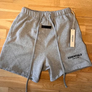 Grå shorts från Essentials Fear of God - Snygga grå shorts från Essentials Fear of God med elastisk midja och långa snören framtill. Klassisk och enkel design med logga tryckt på benet. Perfekta för en avslappnad stil.