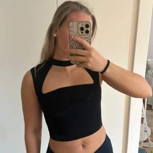 Svart croppad topp med cut-out - Snygg svart croppad topp med unik cut-out detalj vid bröstet. Toppen är ärmlös och har en tight passform som framhäver figuren. Perfekt för att skapa en trendig och stilren look🖤