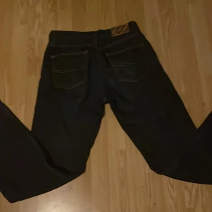Mörkblå jeansbyxor ifrån mcgordon - Säljer ett par klassiska mörkblå jeans med raka ben och kontrastsömmar. Byxorna har fem fickor och stängs med dragkedja och knapp. Perfekta för dig som gillar en tidlös stil.