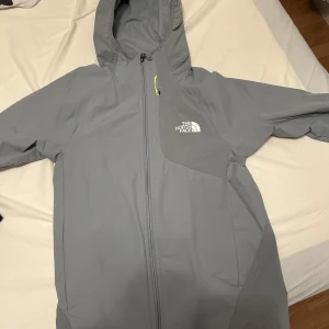 Ljusgrå vindjacka från The North Face - Säljer en ljusgrå vindjacka från The North Face med huva och hel dragkedja framtill. Jackan har en stilren design med logga på bröstet och är perfekt för blåsiga dagar. Passar dig som vill ha en lätt och funktionell jacka.