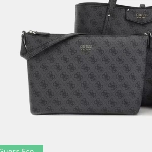 Svart axelväska från Guess - Snygg svart axelväska från Guess med diskret logomönster över hela väskan och silvriga detaljer. Väskan har justerbar axelrem och dragkedja upptill. Perfekt för att bära dina viktigaste saker med stil.