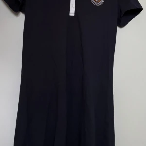 Marinblå pikéklänning från Marine Classics - Säljer en marinblå pikéklänning från Marine Classics med vit krage.  Klänningen har korta ärmar och ett broderat märke på bröstet. Perfekt för en sportig och stilren look.