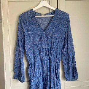 Blå mönstrad playsuit med v-ringning - Säljer en blå playsuit med vitt cirkelmönster och v-ringad hals. Klänningen har lång ärm och resår i midjan för en smickrande passform. Perfekt för vår och sommar! Superfin till sneakers eller sandaler.