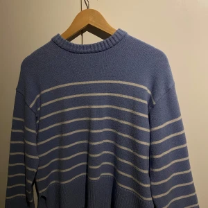 Blåvit randig stickad tröja - Säljer en blå stickad tröja med vita horisontella ränder. Tröjan har rund halsringning, långa ärmar och bred ribbad mudd nedtill samt vid ärmslut. Perfekt för en avslappnad och stilren look.