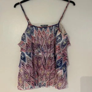 Mönstrad volangig linnetopp från Lindex - Färgglad linnetopp från Lindex med smala axelband och flera volanglager. Toppen har ett abstrakt mönster i blått, rosa, vitt och gult samt dekorativa pärlor längs halsringningen. Perfekt för varma dagar.