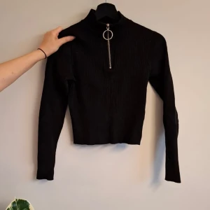 Svart ribbad tröja från H&M - Säljer en svart ribbad croppad tröja från H&M med hög krage och silverfärgad dragkedja framtill. Tröjan har långa ärmar och en tight passform, perfekt till många olika outfits.