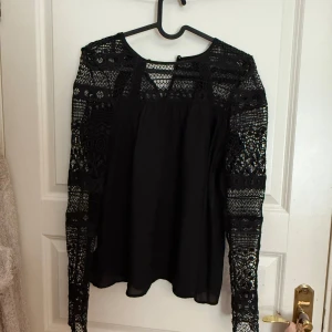 Svart blus med spetsdetaljer - Säljer en svart blus med långa ärmar och vackra spetsdetaljer på axlar, ärmar och över bröstet. Blusen har rund halsringning och ett luftigt fall. Perfekt för dig som gillar stilrena plagg med det lilla extra.