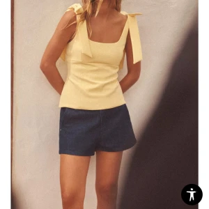 Gul linnetopp med knytband - Snygg ljusgul linnetopp med breda axelband som knyts i rosetter på axlarna. Toppen har rak passform och fyrkantig ringning, perfekt för varma dagar.