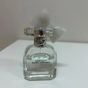 Perfekt Eau de Parfum från Marc Jacobs - En elegant parfymflaska i genomskinligt glas med silverdetaljer och en unik vit dekor på locket. Flaskan har en modern, stilren form och texten 'Perfect Marc Jacobs' på framsidan. Perfekt för dig som gillar exklusiva dofter och snygg design. Det är ungefär 23 ml kvar av 30 ml