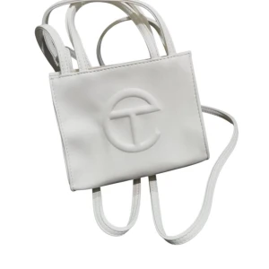Vit axelväska från Telfar - Säljer en stilren vit axelväska från Telfar med präglad logotyp på framsidan. Väskan har både korta handtag och en lång axelrem, perfekt för att bära på olika sätt. Tillverkad i syntetmaterial och har en minimalistisk design.