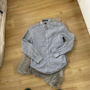 Randig skjorta från GANT - Säljer en klassisk blåvit randig skjorta från GANT i modellen West Coast Oxford. Skjortan har lång ärm, knappar framtill och broderad logga på bröstfickan. Tillverkad i bomull och passar perfekt till en stilren look.