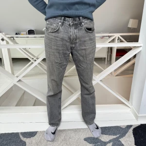 Grå raka jeans - Säljer ett par klassiska grå jeans med raka ben och fem fickor. Jeansen har normal passform och silverfärgade nitar och knappar. Perfekta för en stilren vardagslook.