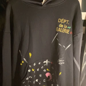 Svart hoodie från Gallery Dept med färgglada detaljer - Svart hoodie från Gallery Dept med trycket 'DÉPT. de la GALERIE' i gult, grönt och rött på bröstet. Unik handmålad känsla med färgprickar och mönster både fram och bak. Mjuk bomullskvalitet och avslappnad passform.