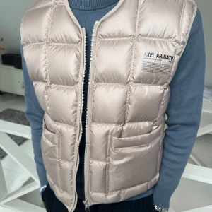 Beige dunväst från Axel Arigato - Stilren beige dunväst från Axel Arigato med quiltad design och dragkedja framtill. Perfekt för lager-på-lager och enkel att matcha med olika outfits.  Storlek XS men väldigt stor i storlek( mer som S/M) 