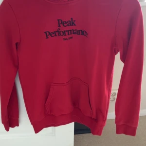 Hoodie - Säljer en röd hoodie från Peak Performance med svart logga på bröstet. Tröjan har lång ärm, magficka och ribbade muddar. Perfekt för en avslappnad stil. Den är storlek 160