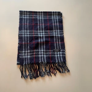 Burberry halsduk - Säljer en klassisk rutig halsduk från Burberry i 100% kashmir. Halsduken har en mörkblå bas med vita och röda rutor samt fransar i ändarna. Perfekt accessoar för att lyfta din outfit.