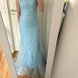 Ljusblå balklänning  - Säljer en ljusblå långklänning med tunna axelband och vackra blommiga broderier. Klänningen har en figurnära passform, tyll i kjolen och snörning i ryggen. Perfekt för bal eller festliga tillfällen.