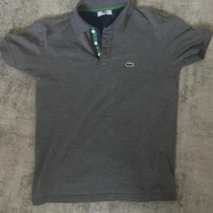 Grå piké t-shirt från Lacoste - Säljer en klassisk grå piké t-shirt från Lacoste med korta ärmar och knappslå framtill. Ikoniska krokodilloggan broderad på bröstet och gröna detaljer på insidan av kragen. Perfekt för en stilren look.
