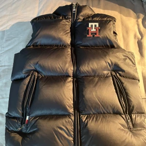 Limited Edition Tommy Hilfiger väst - Säljer en svart dunväst från Tommy Hilfiger med TH-logga på bröstet och broderad HILFIGER-text på kragen. Västen har två snedställda fickor med dragkedja och hög krage. Perfekt för lager-på-lager under kyliga dagar.