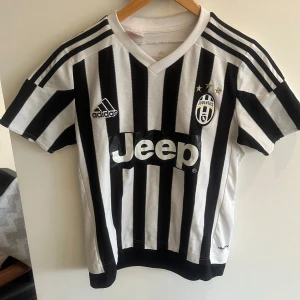 Juventus fotbollströja (2015-16), stl 164 - Juventus matchtröja från Adidas med klassiska svarta och vita ränder, Jeep-logga på bröstet och klubbmärke med tre stjärnor. Kortärmad modell med V-ringning och broderade detaljer. Perfekt för fotbollsfans!