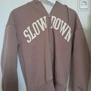 Brun hoodie med dragkedja  - Mysig brun hoodie från Gina Tricot med vit text 'SLOW DOWN' på bröstet. Tröjan har huva, långa ärmar och dragkedja framtill. Perfekt för en avslappnad stil och enkel att matcha med jeans eller leggings. Inget fel men kan diskutera priset🦄🎉