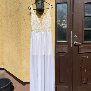 Vit balklänning med spets och djup v-ringning - Elegant vit balklänning med spets upptill och djup v-ringning. Klänningen har ärmlös design och flowy kjol i tunt material. Perfekt för speciella tillfällen och ger en romantisk känsla.