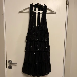 Svart glittrig volangklänning från SHEIN - Supersnygg svart kortklänning från SHEIN med glittriga paljetter och flera volanglager. Djup v-ringning och öppen rygg med knyt i nacken. Perfekt för dig som vill sticka ut på festen!