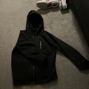 Svart hoodiejacka från Sail Racing - Svart hoodiejacka från Sail Racing med dragkedja framtill och huva. Jackan har en enkel design med logga på bröstet och är perfekt för en avslappnad stil.