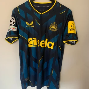 Newcastle United bortatröja Isak 14 - Snygg mörkblå Newcastle United bortatröja med gula detaljer och tryck 'Isak 14' på ryggen. Tröjan har korta ärmar, geometriskt mönster i blått och gult samt Champions League-märke på ärmen. Perfekt för fotbollsfans! Den är ifrån 2023-24 säsongen. 