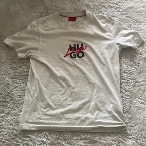 Vit t-shirt från Hugo - Säljer en vit t-shirt från Hugo med tryckt logga i svart och rött på bröstet. Klassisk passform och rund halsringning. Perfekt för en stilren och enkel look.