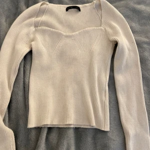 Vit ribbad långärmad topp - Säljer en vit ribbad långärmad topp med unik skärning vid halsen och markerade sömmar framtill. Toppen har en figurnära passform och är tillverkad i ett stretchigt material som sitter snyggt på kroppen.