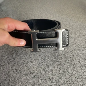 Svart bälte med H-spänne - Säljer ett svart skärp i skinn med ett stort, ikoniskt H-spänne i silverfärgad metall. Bältet har vit kontrastsöm längs kanterna och en stilren, klassisk design som verkligen sticker ut. 