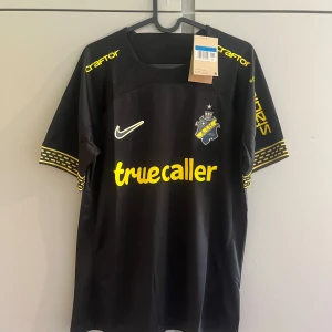 AIK svart matchtröja Nike 2024 - AIK:s officiella svarta matchtröja från Nike med gula detaljer och tryck. Tröjan har klubbmärke, sponsorloggor och unika gula mönster på ärmsluten. Tillverkad i lätt och ventilerande funktionsmaterial, perfekt för fotboll eller supporterliv.