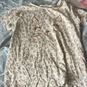 Beige leopardmönstrad t-shirt H&M - Söt beige t-shirt från H&M med korta ärmar och leopardmönster över hela plagget. Framsidan har ett glittrigt kattansikte tryckt i guld. Tillverkad i mjuk bomull, perfekt för en lekfull och trendig look.