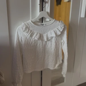 Vit blus med volang från Kappahl - Supersöt vit blus från Kappahl i storlek 146/152. Blusen har lång ärm, strukturmönster och en bred volang runt halsringningen som ger en romantisk vibe. Perfekt för dig som gillar en stilren men ändå lekfull look.💗 aldrig använd 💗