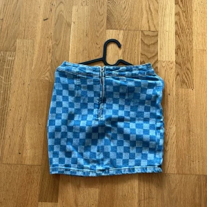 Blå rutig jeanskjol med slits - Snygg kort jeanskjol i blått med rutigt mönster. Kjolen har dragkedja bak och en liten slits framtill för extra edge. Perfekt till sneakers eller boots för en cool look.