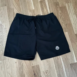Svarta shorts från Moncler med snörning - Svarta shorts från Moncler i nylon med elastisk midja och snörning. Klassisk Moncler-logga på ena benet. Perfekta för sommaren och chill dagar. Enkel och stilren design som funkar till det mesta. Storleken står att det är xxl men fattar inte riktigt de för de passar meg perfekt som är 175cm