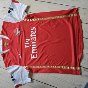 Arsenal tröja - Arsenal hemmatröja med Walcott 32 på ryggen. Röd kropp med vita korta ärmar, guldiga detaljer och klubbmärke på bröstet. Nike FIT DRY-material som andas, tryckt sponsor och Premier League-patch på ärmarna. Perfekt för dig som älskar Arsenal och klassiska tröjor. 