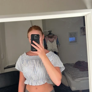 Randig offshoulder croppad topp FB Sister - Gullig croppad topp från FB Sister i vitt och ljusblått med ränder. Offshoulder-modell med smock upptill, volangkant nertill och söt rosett i ryggen. 