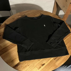 Svart sweatshirt från Polo Ralph Lauren - En stilren svart sweatshirt från Polo Ralph Lauren med klassisk rund hals och diskret broderad logga på bröstet. Tröjan har långa ärmar och breda muddar vid ärmslut och nederkant. Perfekt för en clean och avslappnad look.