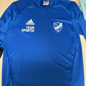 Blå IFK Eskilstuna fotbollströja Adidas - Snygg blå fotbollströja från Adidas med IFK Eskilstuna-logga och TEAM SPORTIA-tryck på bröstet. Tröjan har lång ärm och klassisk rund hals. Perfekt för träning eller match. Tillverkad i lätt och funktionellt material.