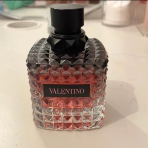 Valentino parfym med nitar - Snygg parfym från Valentino i en genomskinlig glasflaska med nitar och svart lock. Flaskan har en cool rutstruktur och rödrosa vätska inuti. Märket står tydligt på framsidan. Perfekt för dig som gillar lyxiga och trendiga dofter.