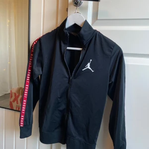 Jordan tröja/barn  - Svart barntröja Jordan med vit logga på bröstet och röda detaljer längs ärmarna där det står 'JORDAN'. Jackan har hel dragkedja framtill och hög krage. Perfekt för dig som gillar sportig och clean stil. Passar barn i 9-10 år skulle jag säga. 