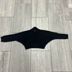 Svart glittrig kofta från Gina Tricot - Snygg svart glittrig kofta från Gina Tricot i storlek S. Modellen är kort och öppen framtill med lång ärm. Materialet är stickat med diskret glittertråd som ger en extra touch. Perfekt att slänga över en topp för en cool look.