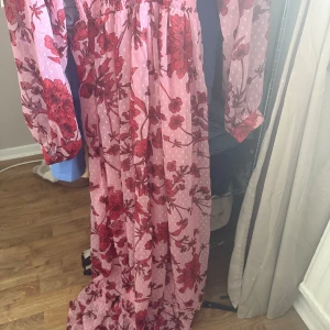 Rosa blommig maxiklänning Boohoo - Supersöt långklänning från Boohoo i rosa med röda blommor och små vita prickar. Klänningen har v-ringning, långa ärmar med resår vid handlederna och smock i midjan för en snygg passform. Perfekt för dig som vill sticka ut med färg och mönster!